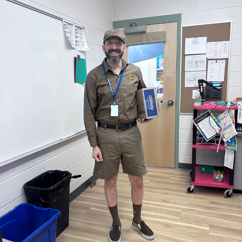 A man dressed as a UPS delivery driver holds a Chewy box in a classroom.