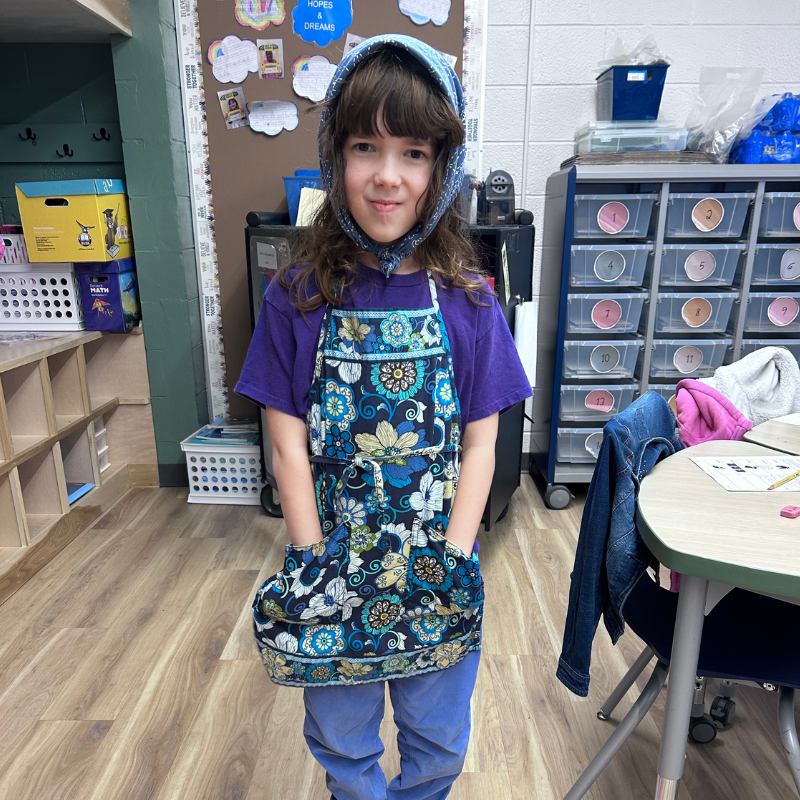 A young person stands in a classroom wearing a patterned apron and a blue headscarf.