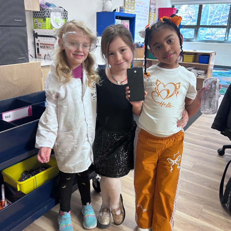 Three young girls dressed in costumes pose together in a classroom.