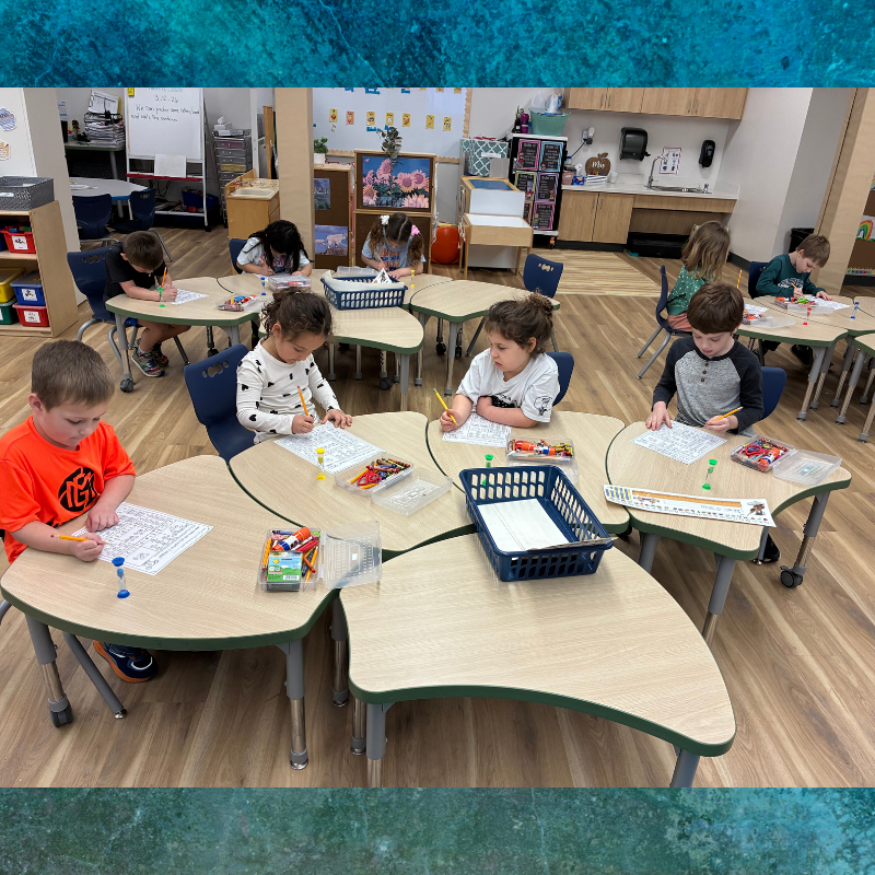 Children are focused on writing and drawing at clover-shaped desks in a bright classroom.