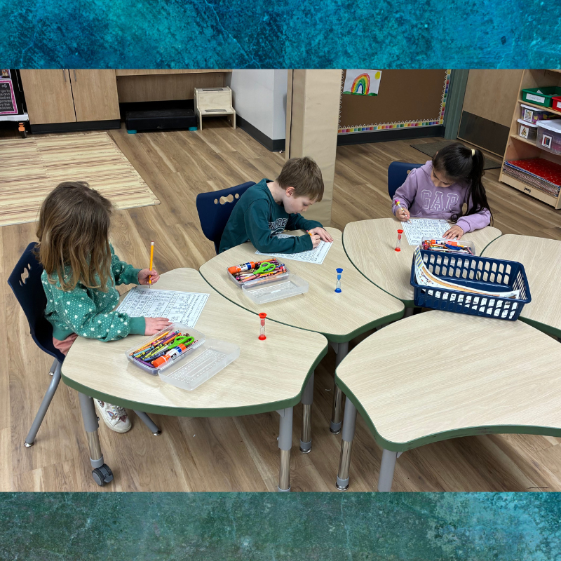 Three children focus on their worksheets at clover-shaped tables in a classroom.