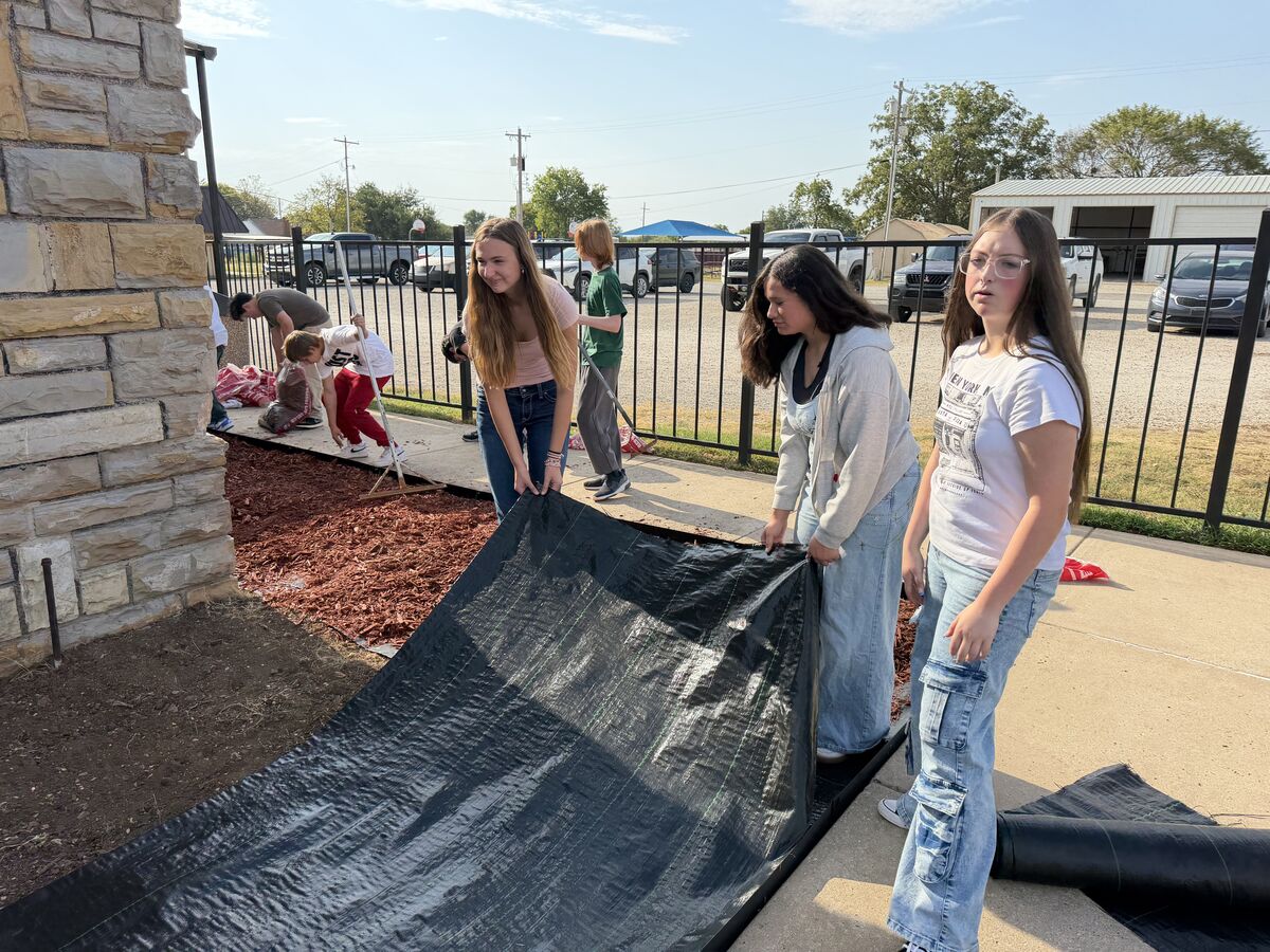 A group of young people are working outdoors, laying down black landscape fabric near a stone wall.