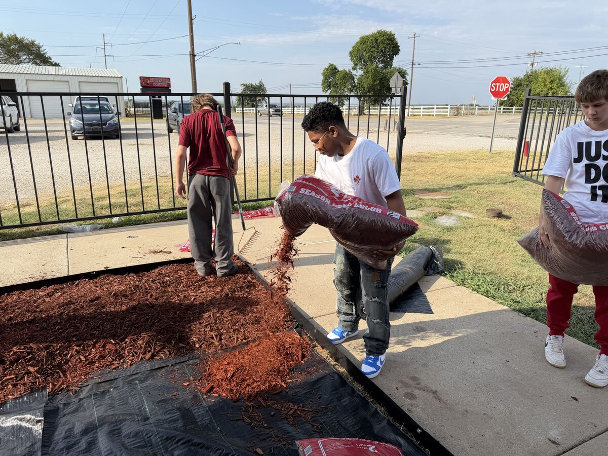 Three young people are spreading red mulch outdoors, with one person pouring from a bag and another raking.