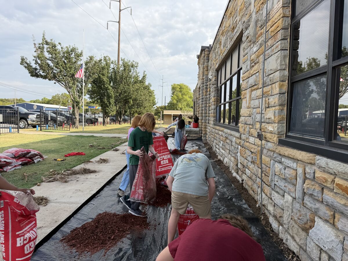 A group of young people are spreading red mulch from bags onto a black landscape fabric next to a stone building.