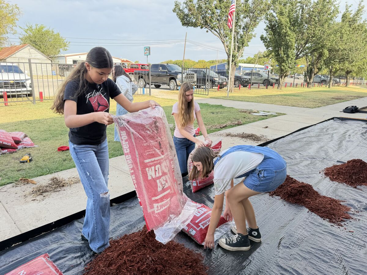 Several young people are spreading red mulch from bags onto a black landscape fabric outdoors.