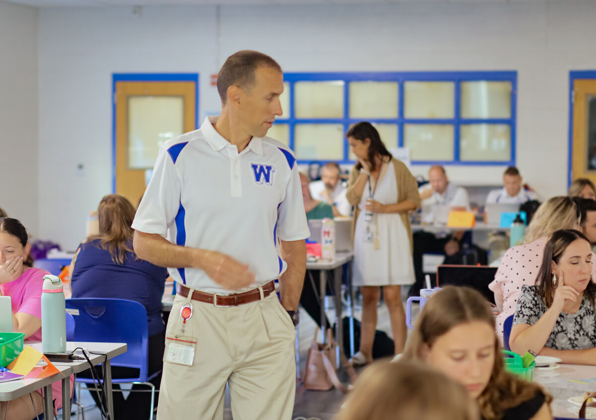 A man in a white polo shirt with a blue 'W' logo stands in a classroom setting.