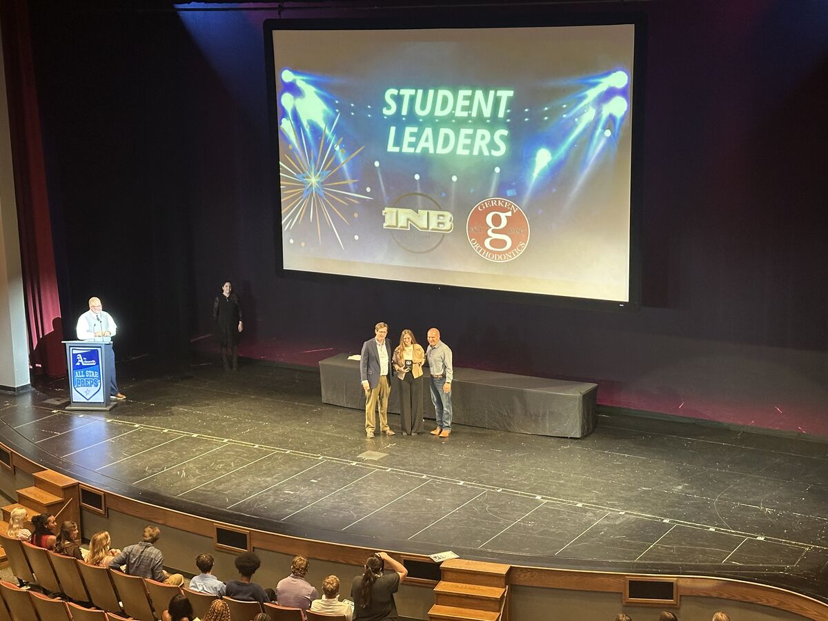 Three individuals stand on a stage in front of a large screen displaying "STUDENT LEADERS" and sponsor logos.