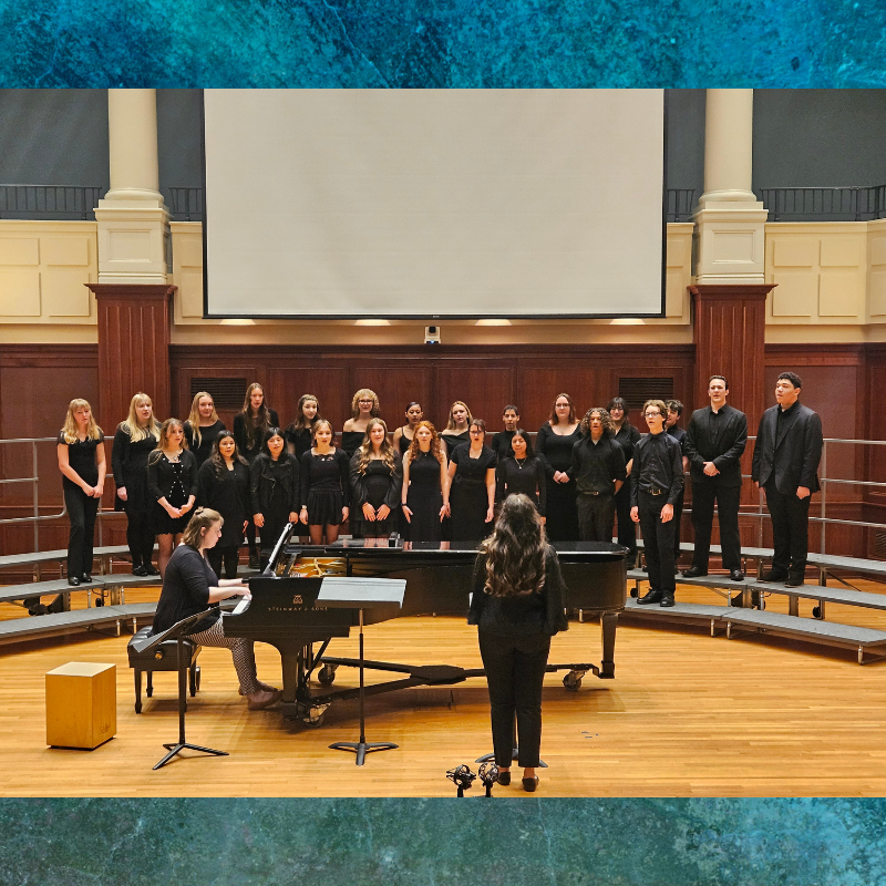 A choir of young people in black attire stands on risers, facing a piano and conductor.