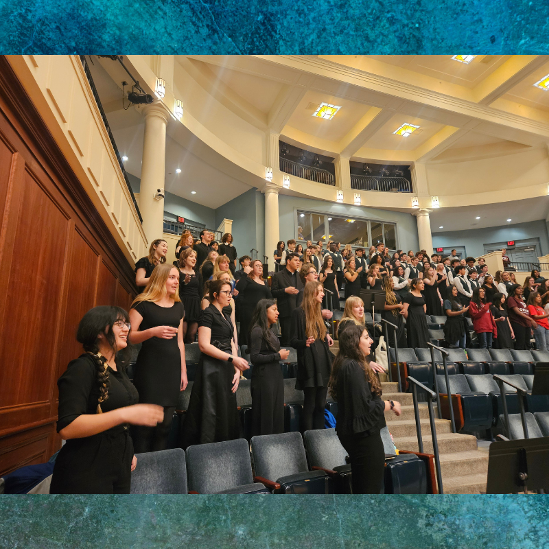 A large choir of young people in black attire stands on tiered seating in an auditorium.