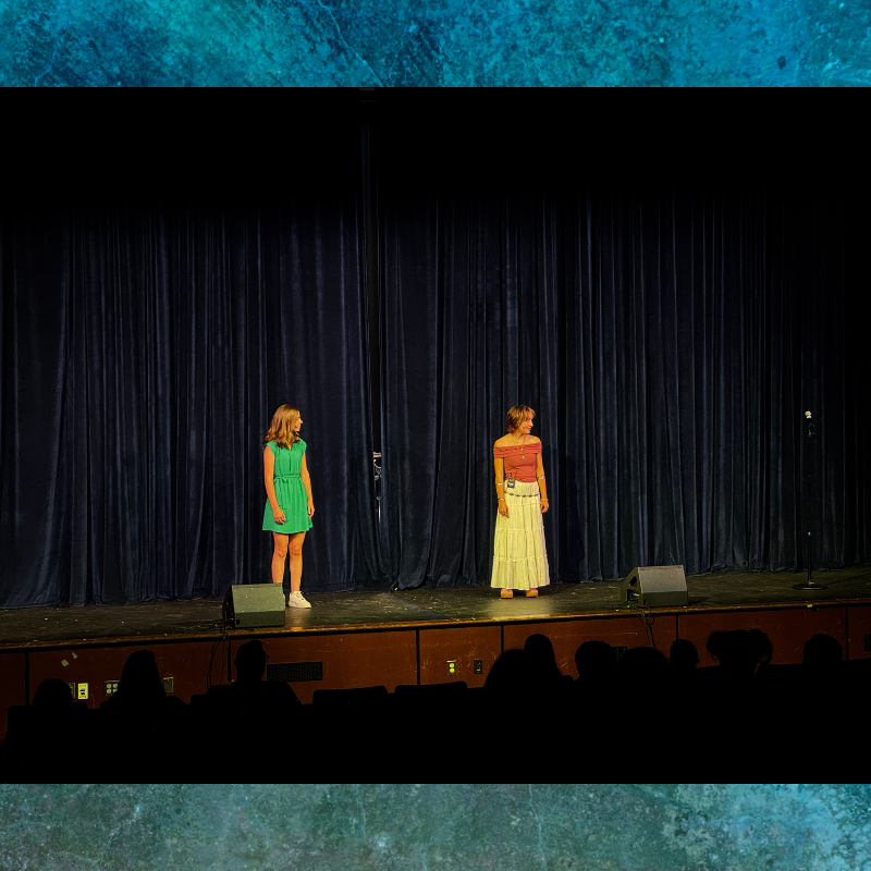Two women stand on a stage in front of a dark blue curtain, facing an audience.