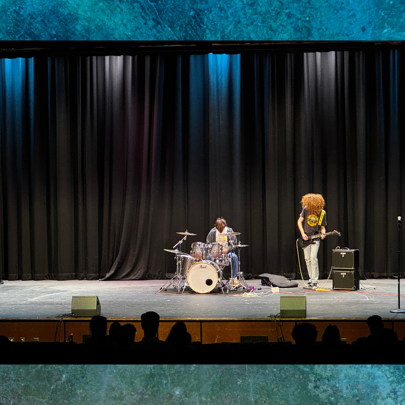 Two musicians perform on a stage with a black curtain backdrop, one playing drums and the other a guitar.