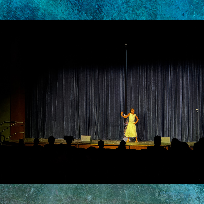 A young performer in a light-colored dress stands on a stage in front of dark blue curtains.