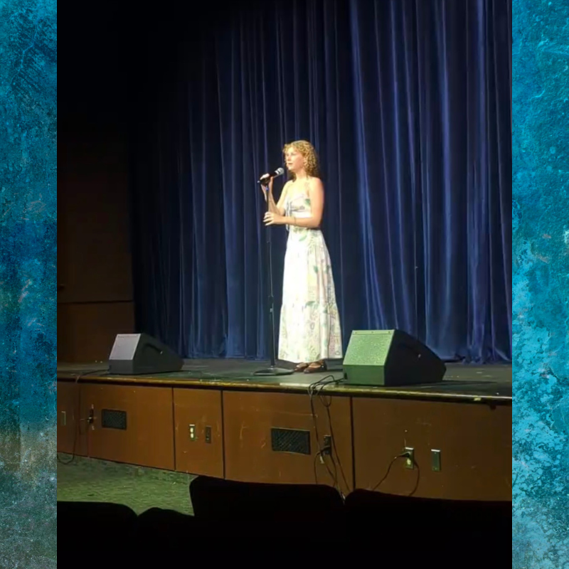 A young woman in a long dress sings into a microphone on a stage with blue curtains.