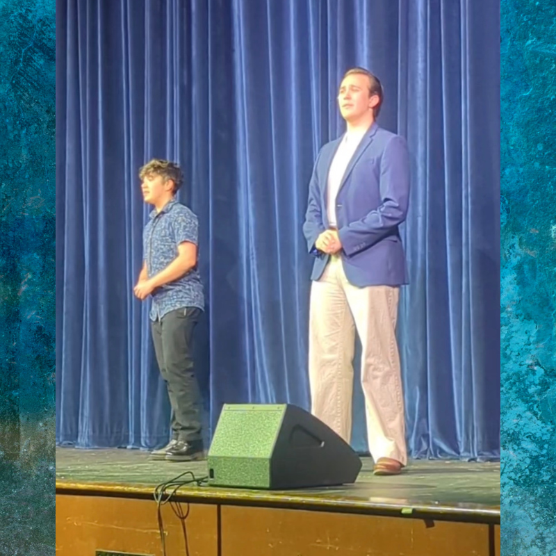 Two young men stand on a stage in front of blue curtains, one in a patterned shirt and the other in a blue blazer.