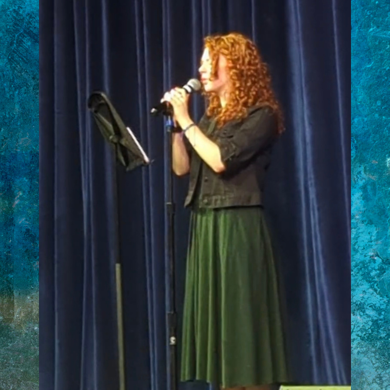A woman with curly red hair sings into a microphone on a stage with blue curtains.