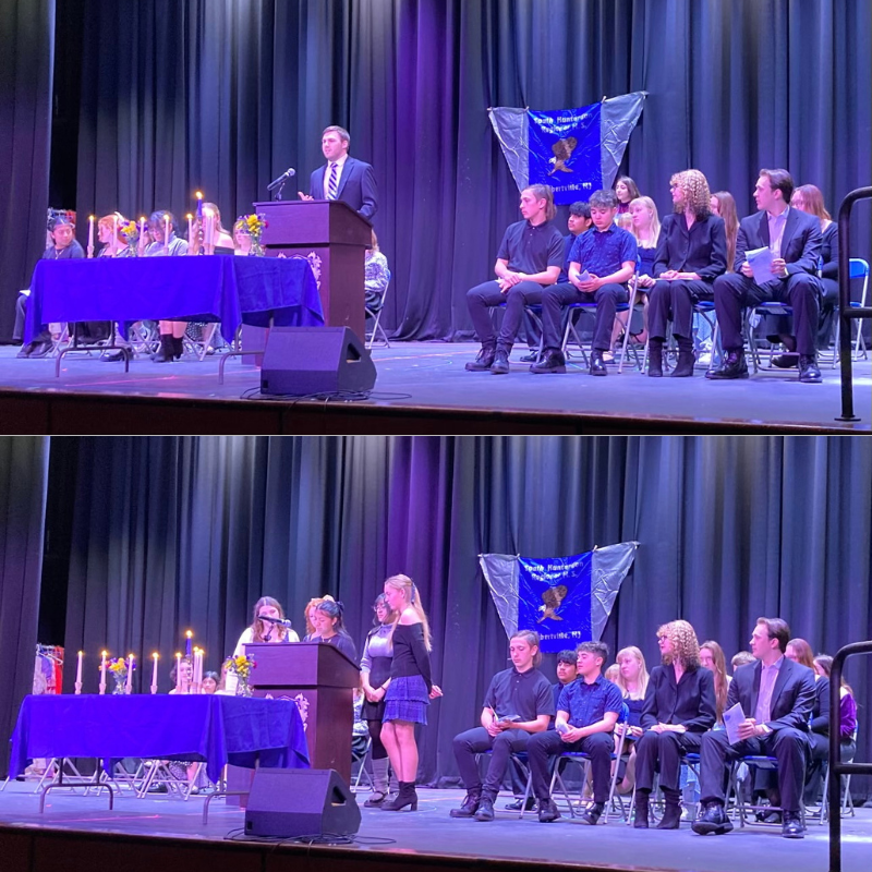 A speaker addresses an audience from a podium on a stage, with students seated behind a table with lit candles.