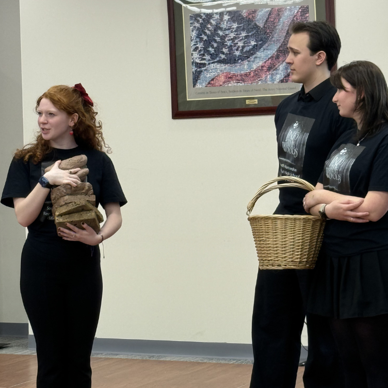 A young woman with red curly hair holds a stack of bread loaves while speaking.