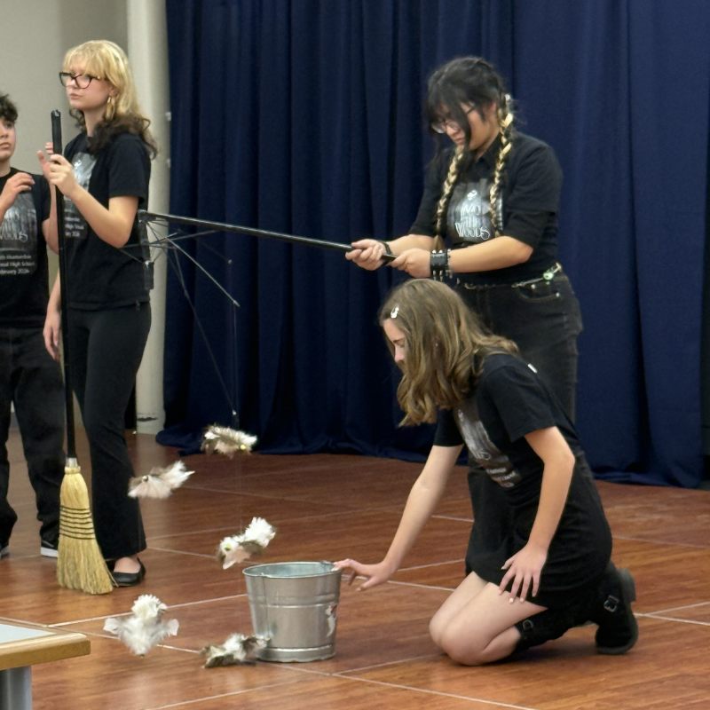 Students in black t-shirts perform a theatrical scene with dangling props and a bucket.
