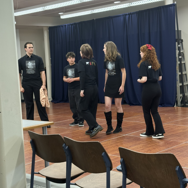 Five people in black t-shirts stand on a wooden stage in front of a blue curtain.