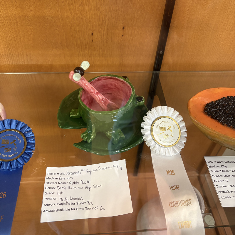 A ceramic sculpture of a green frog with a pink tongue extended, catching a fly, displayed with ribbons.