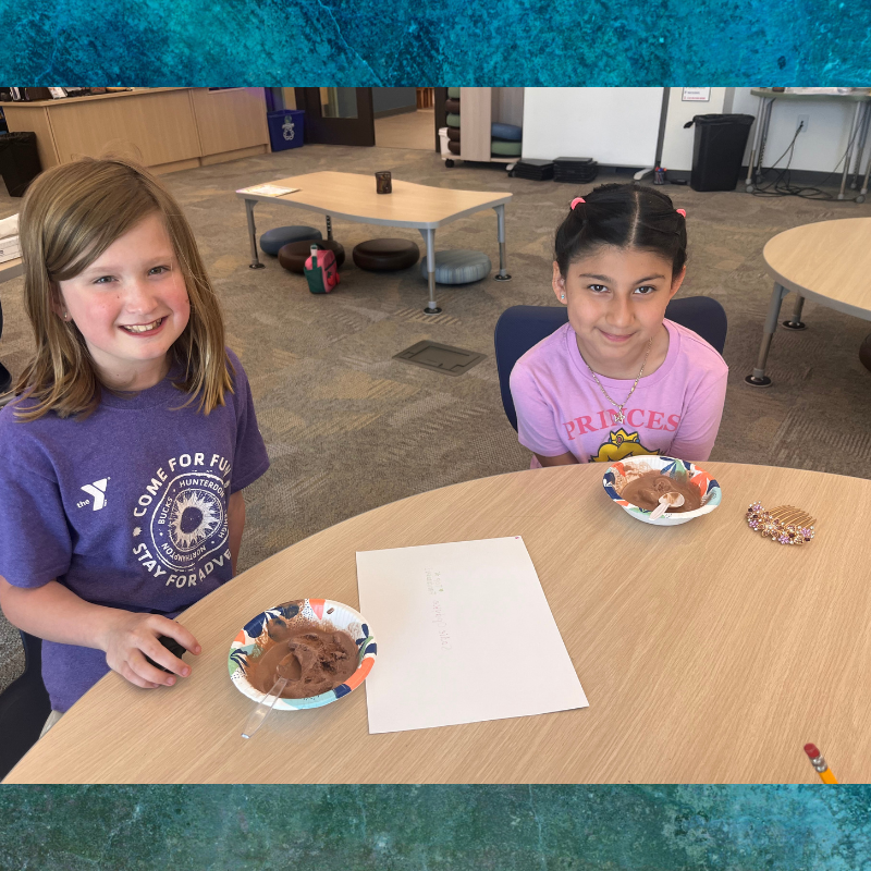 Two young girls smile at the camera while sitting at a table with bowls of chocolate ice cream.