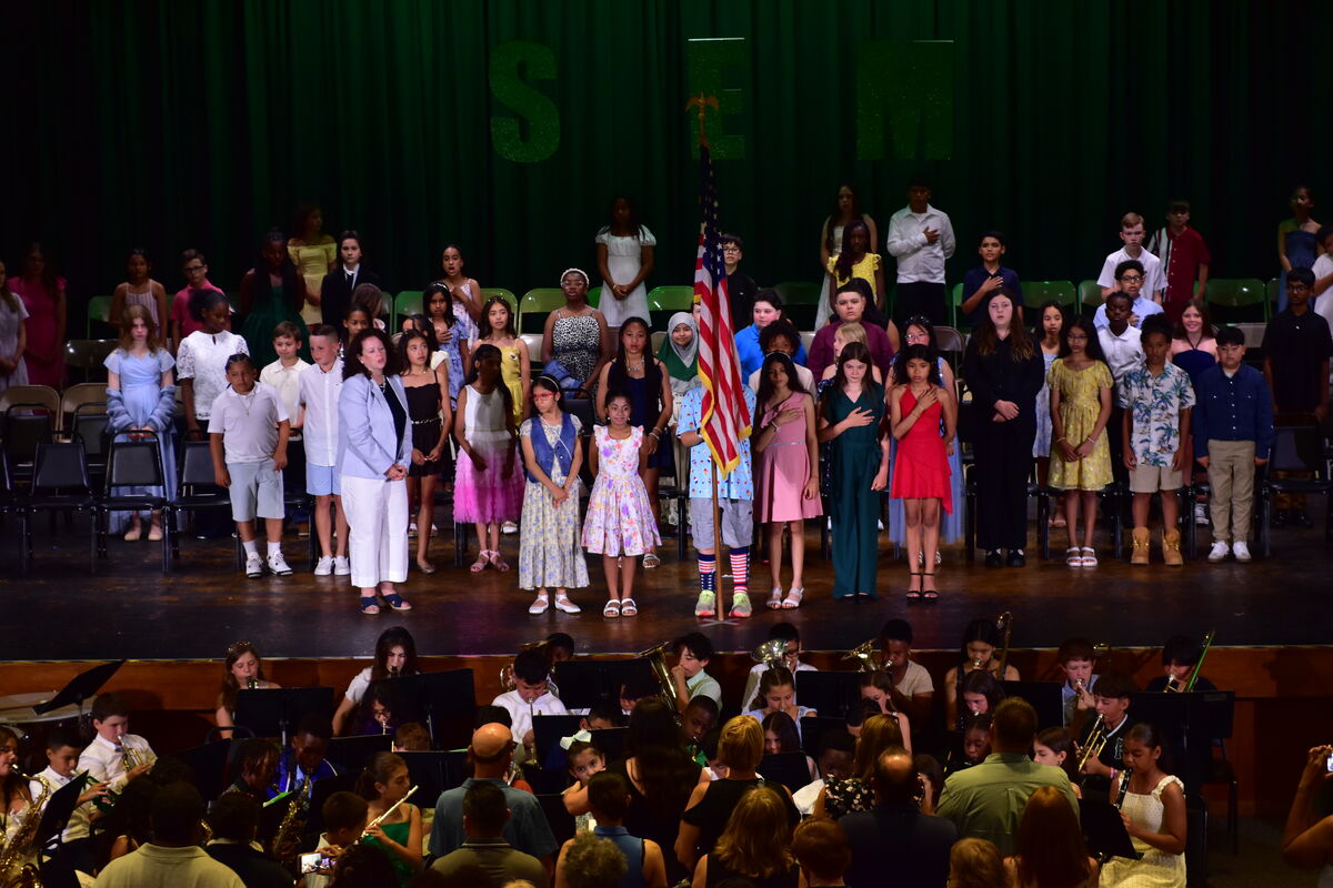A group of students stand on a stage in front of a green curtain, with an American flag displayed.