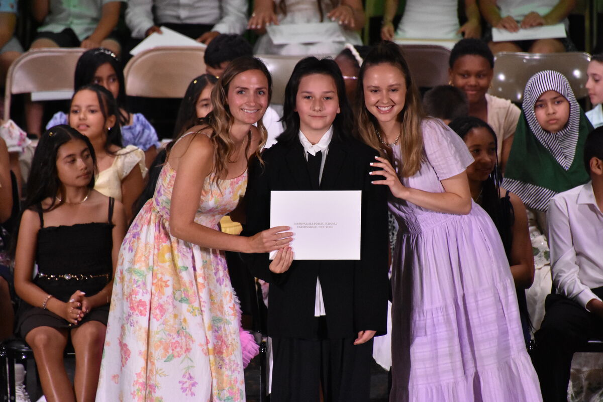 A young person in a black suit holds a certificate with two smiling women beside them at an event.