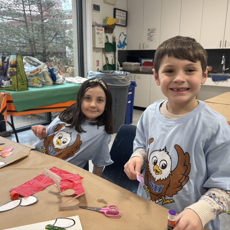 Two children in matching light blue t-shirts with an eagle graphic are engaged in an art project at a table.