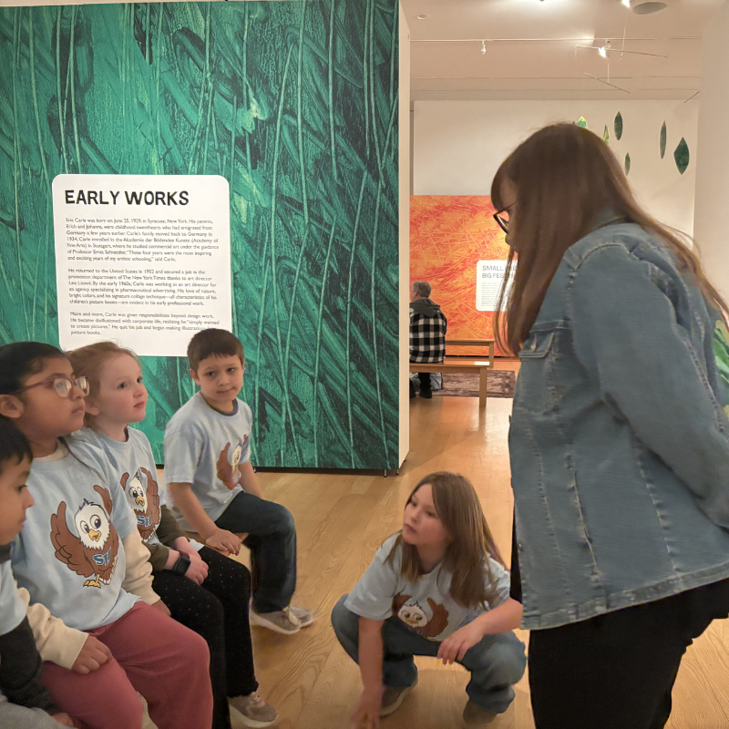 A group of children in matching t-shirts listen attentively to a woman in a denim jacket in an art gallery.