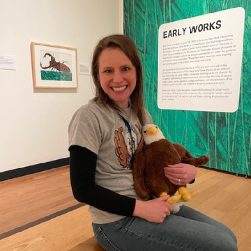 A woman smiles while holding a stuffed eagle in front of an art exhibit.