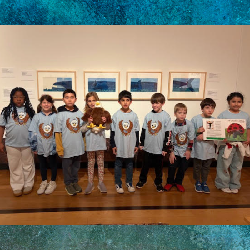 A group of children in matching t-shirts stand in front of framed artwork.