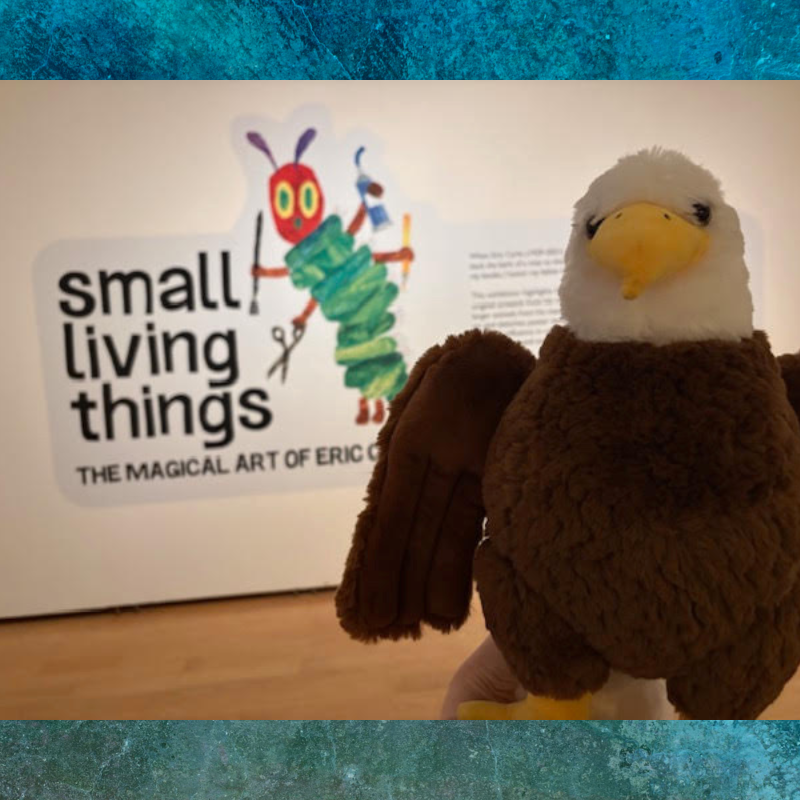 A plush eagle toy stands in front of an exhibit sign that reads 'small living things' and features a colorful caterpillar illustration.