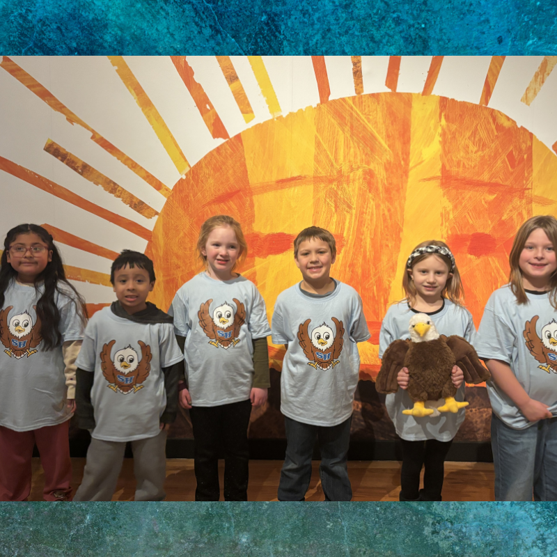 Six children stand in front of a large, vibrant sun mural, all wearing matching light blue t-shirts with an owl logo.
