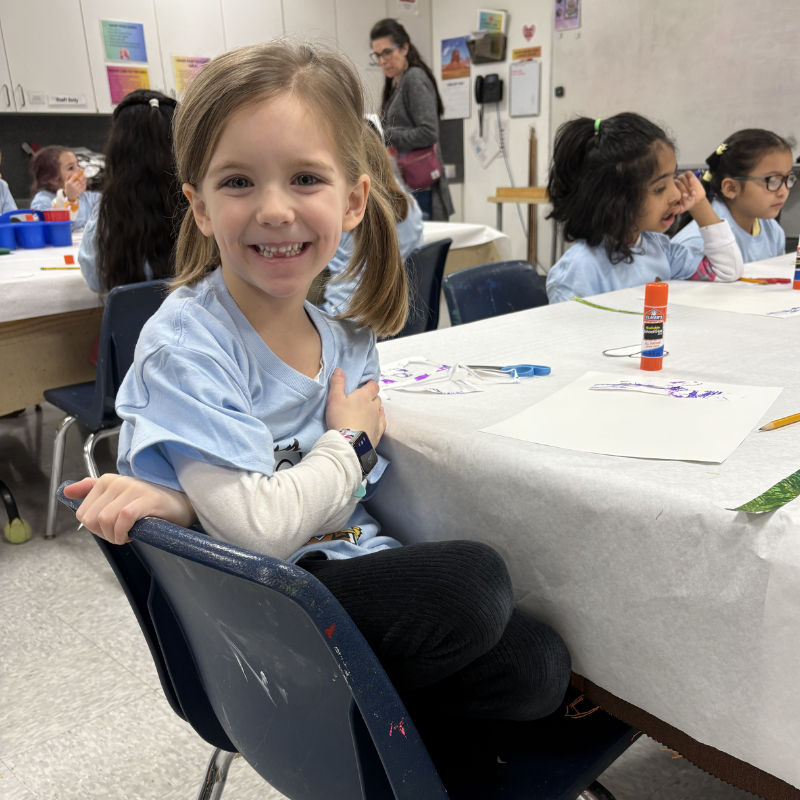 A young girl with pigtails smiles at the camera while sitting at a table in an art class.