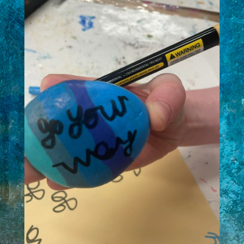 A hand holds a blue and teal painted rock with the words 'go your way' written in black marker.