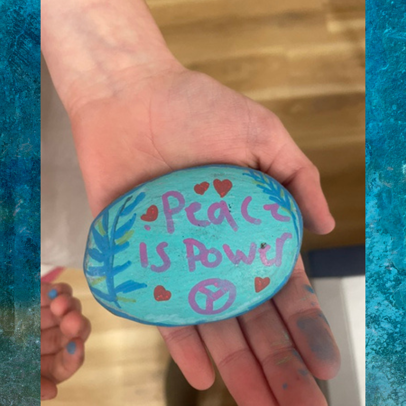 A hand holds a painted rock with the words 'Peace is Power' and a peace symbol.
