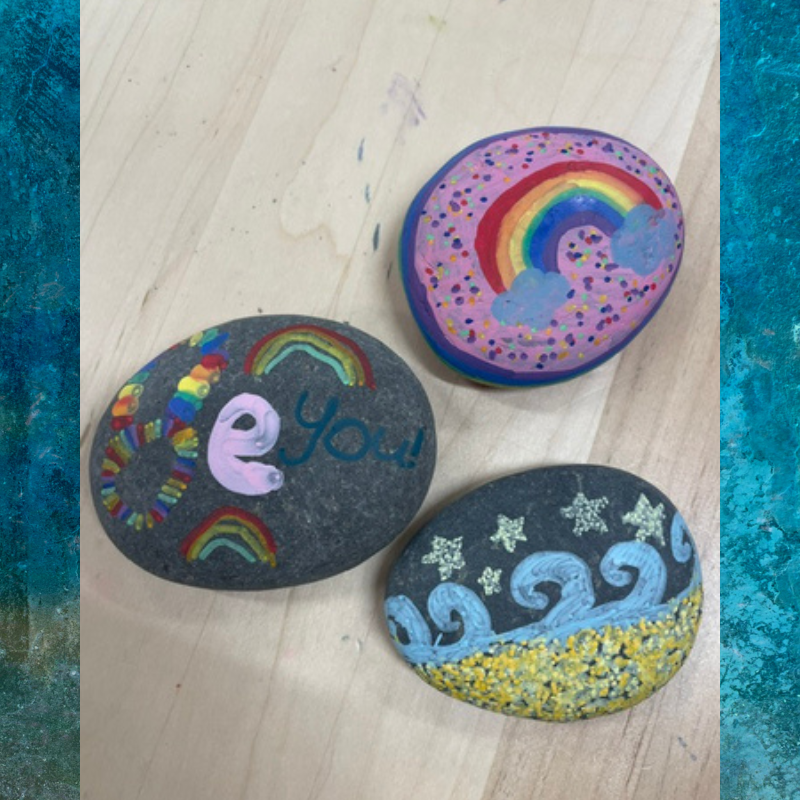 Three painted rocks with colorful designs: a donut with a rainbow, a rock with 'e You!' and rainbows, and a rock with waves and stars.