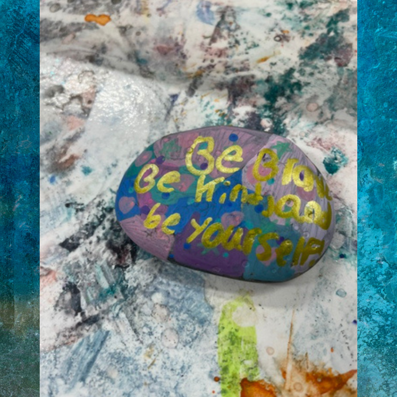 A painted rock with the words 'Be Brave, Be Kind, Be Yourself' in gold lettering.