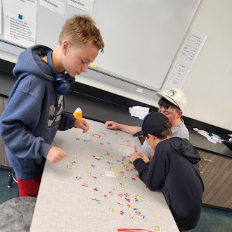 Three students stand around a table in a classroom, sorting through colorful pieces scattered across the surface.