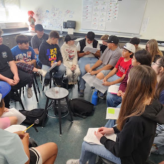 A diverse group of students sits in a circle inside a classroom, reading papers and participating in a group activity.