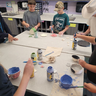 Students gather around tables in a classroom, painting handmade ceramic bowls with colorful paints.