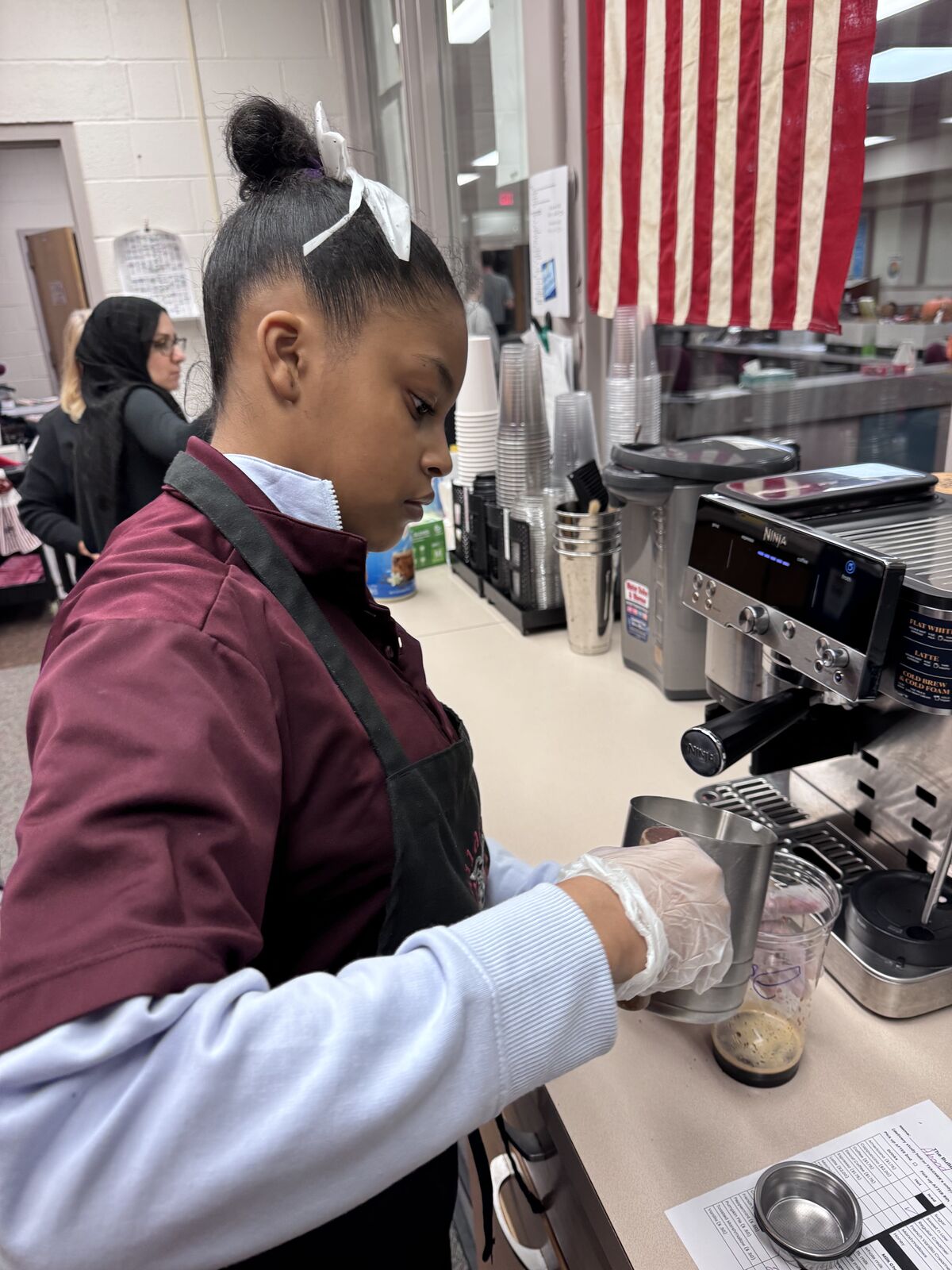 A young person wearing gloves and an apron prepares a beverage in a cafe setting.