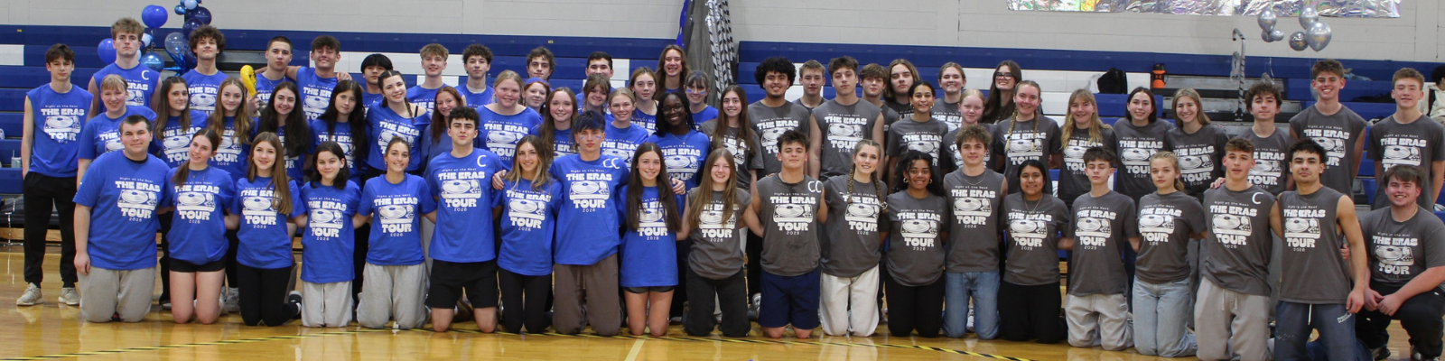 A large group of young people, some in blue shirts and some in gray shirts, pose for a photo in a gymnasium. The shirts have text that reads 'NIGHT AT THE ERAS TOUR 2020'.