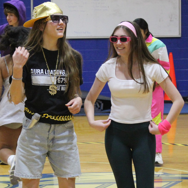 Two young women in retro-inspired outfits smile and pose in a gymnasium.