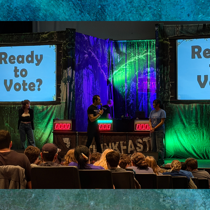 Two presenters stand on a stage with "Ready to Vote?" displayed on screens, in front of a forest-themed backdrop.