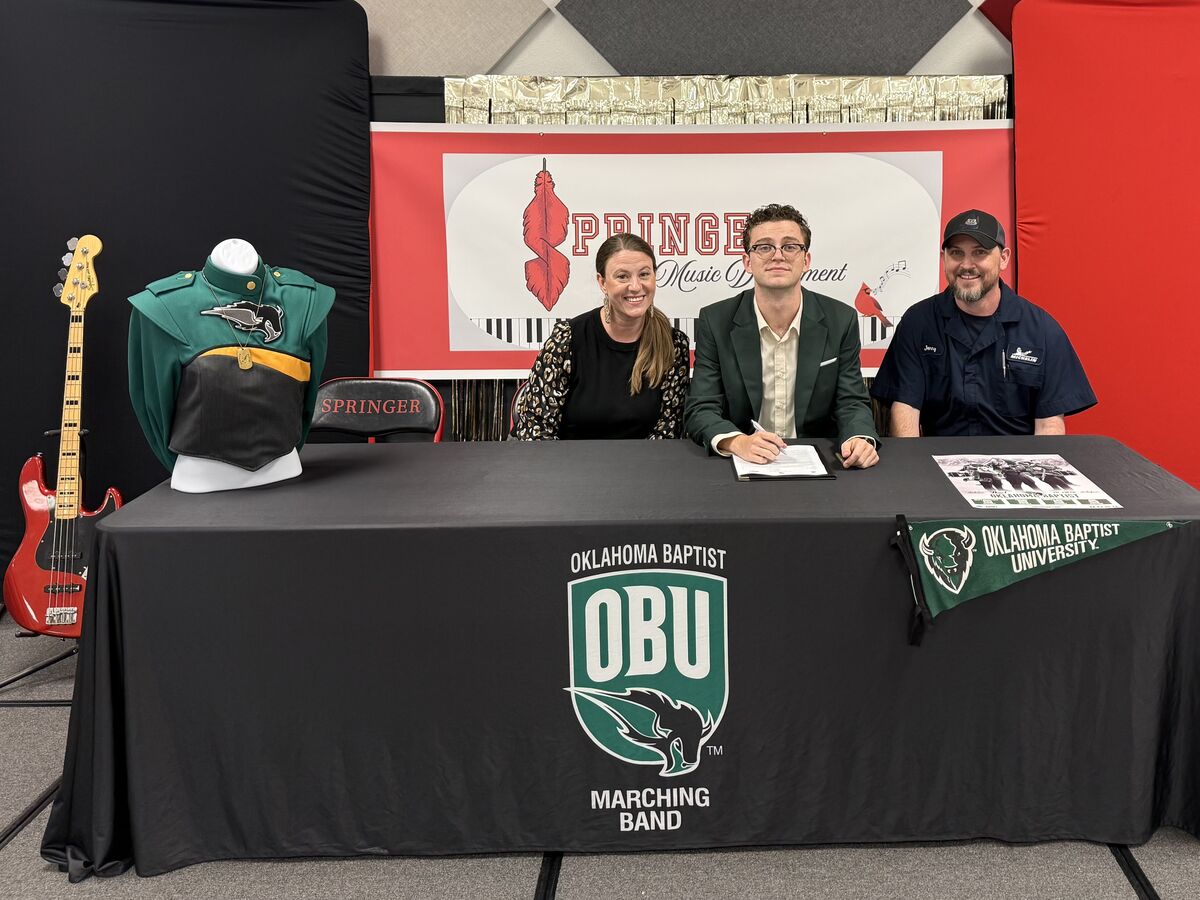 Three people sit at a table with Oklahoma Baptist University Marching Band banners, a uniform on a mannequin, and a bass guitar nearby.