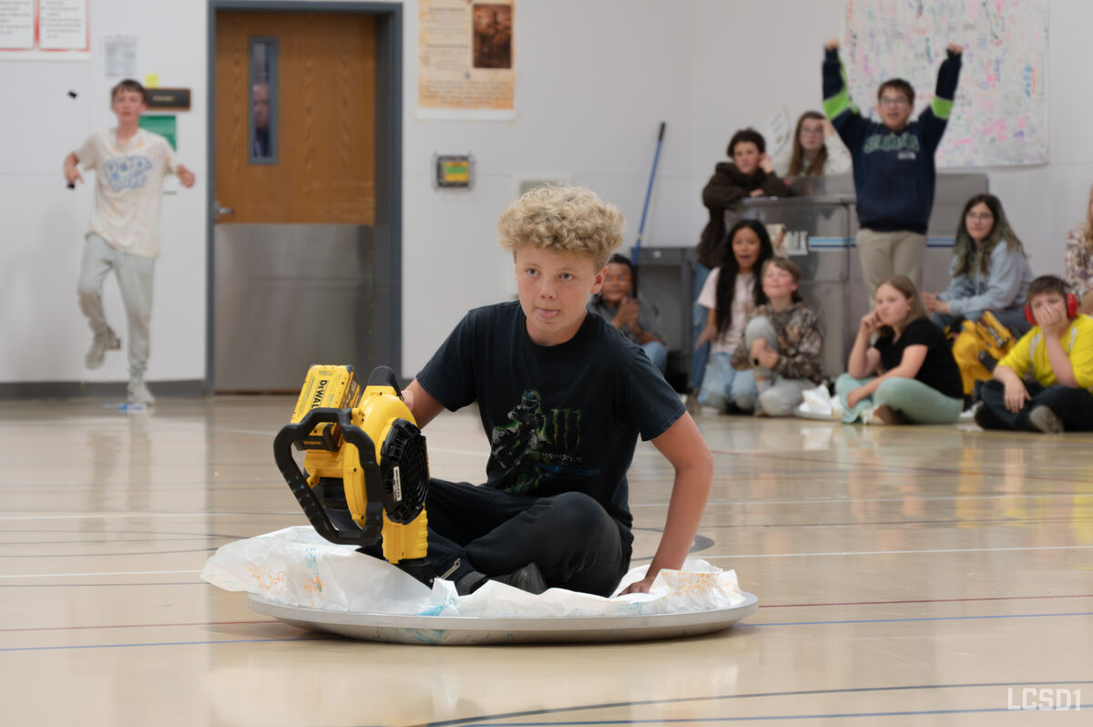 A young person sits on a large platter, holding a yellow leaf blower, in a gymnasium.