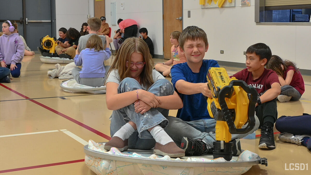 Children sit on the floor in a gymnasium, some with a yellow leaf blower toy.