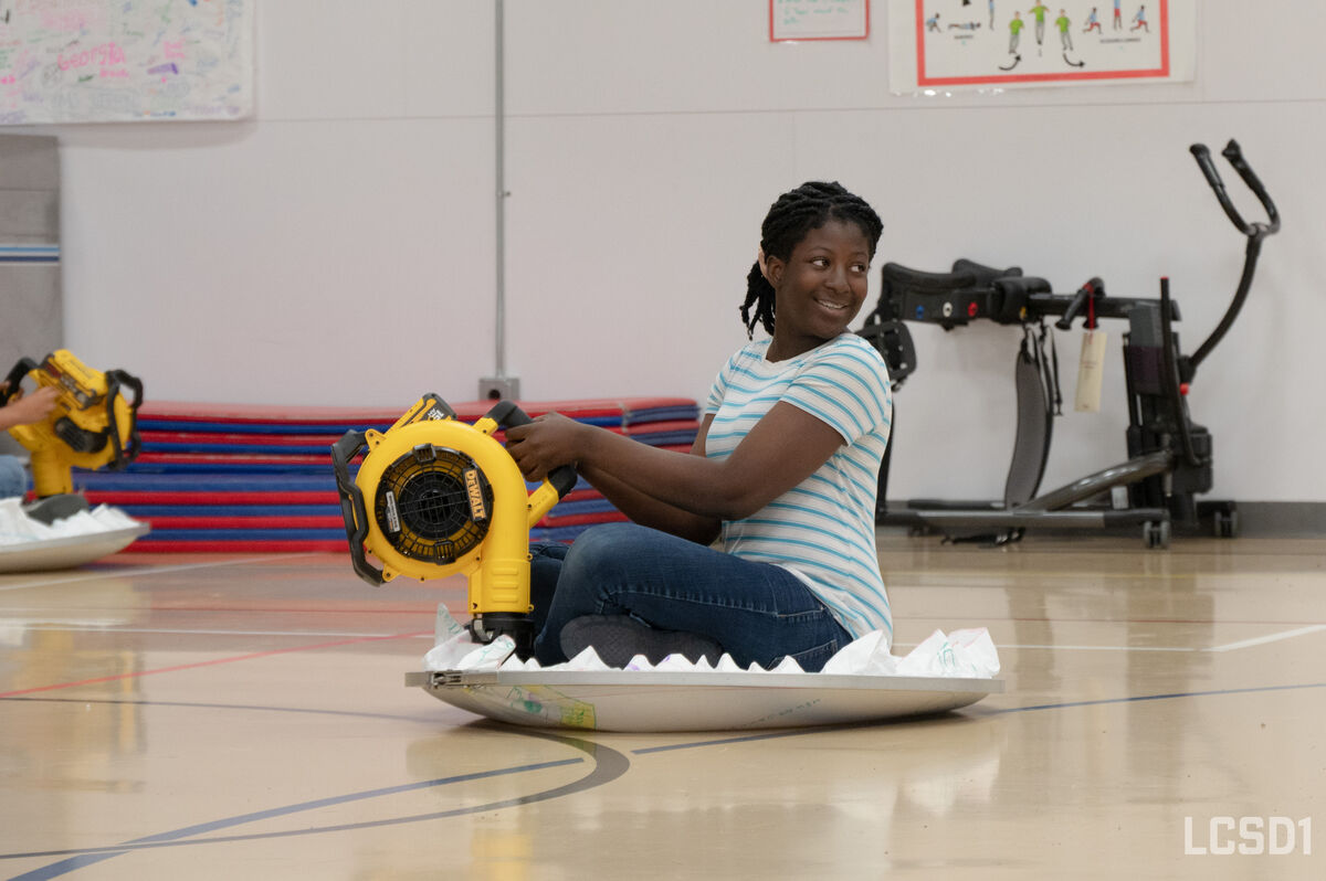 A young person smiles while sitting on a disc and holding a yellow leaf blower in a gymnasium.
