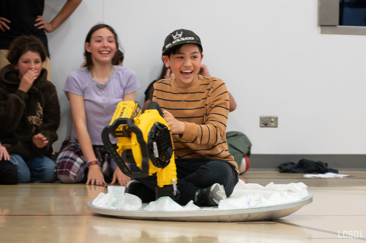 A boy in a striped shirt and baseball cap laughs while holding a yellow leaf blower on a sled.